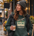 A woman wearing a dark green Sea-Tac themed hoodie with a vintage-style graphic and the words 'FOREVER Sea-Tac', layered under a black puffer vest and a gray beanie, holding a coffee cup on a city street.