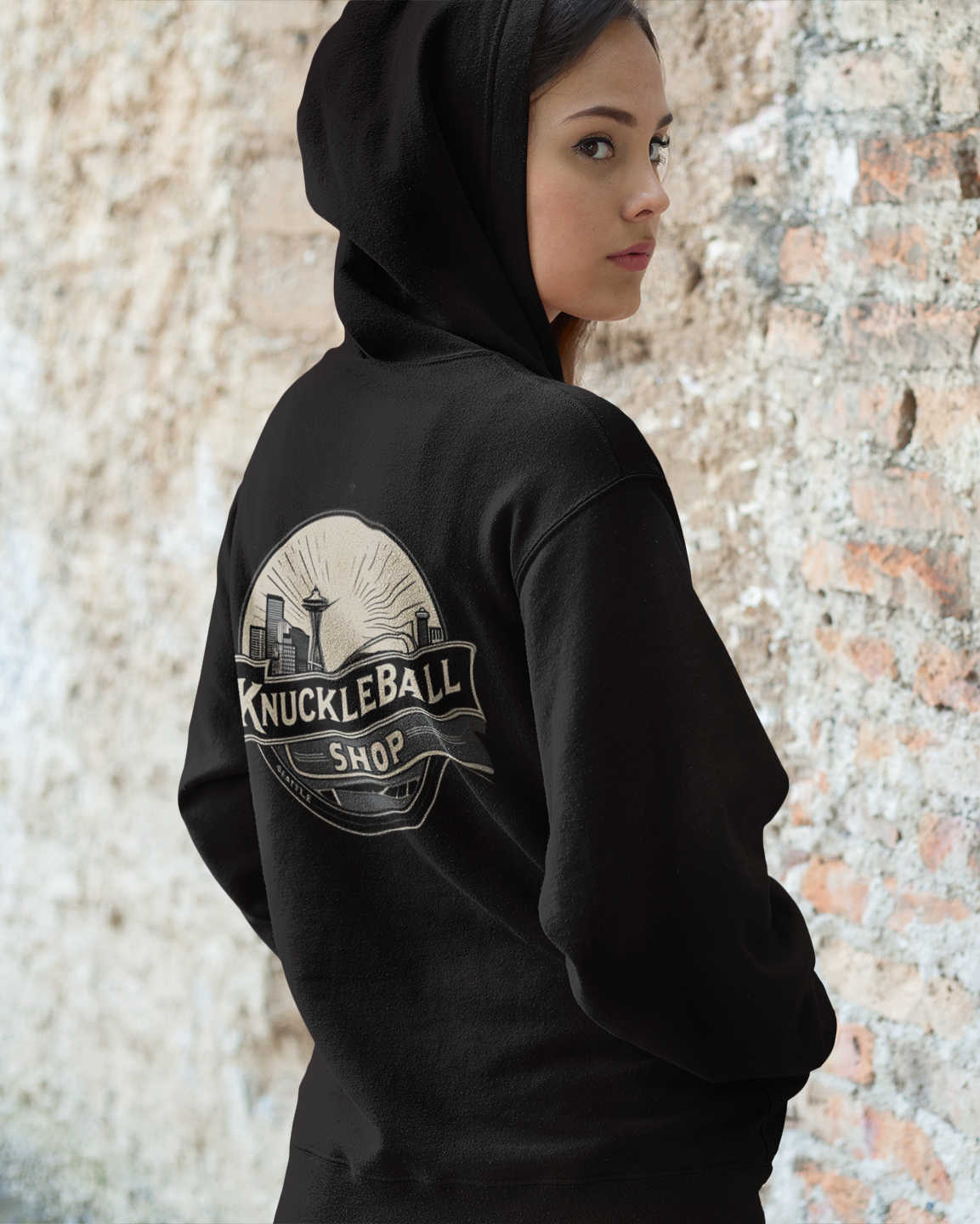 Person wearing a black hoodie with 'Knuckleball Shop' logo against a textured wall.