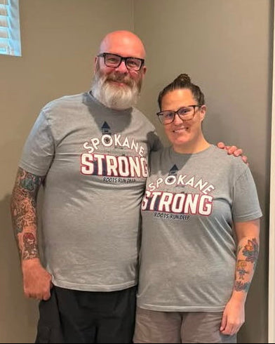 A man and a woman standing indoors wearing matching gray t-shirts with 'SPOKANE STRONG' Roots Run Deep printed on the front.