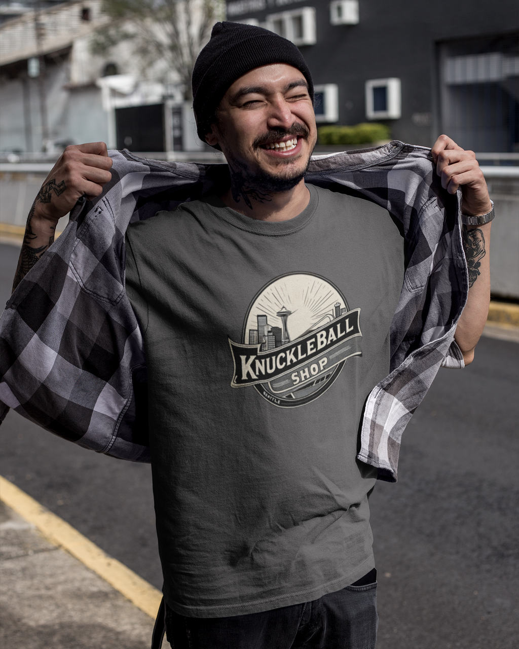 Man wearing a gray t-shirt with 'Knuckleball Shop' logo, holding a checkered shirt.