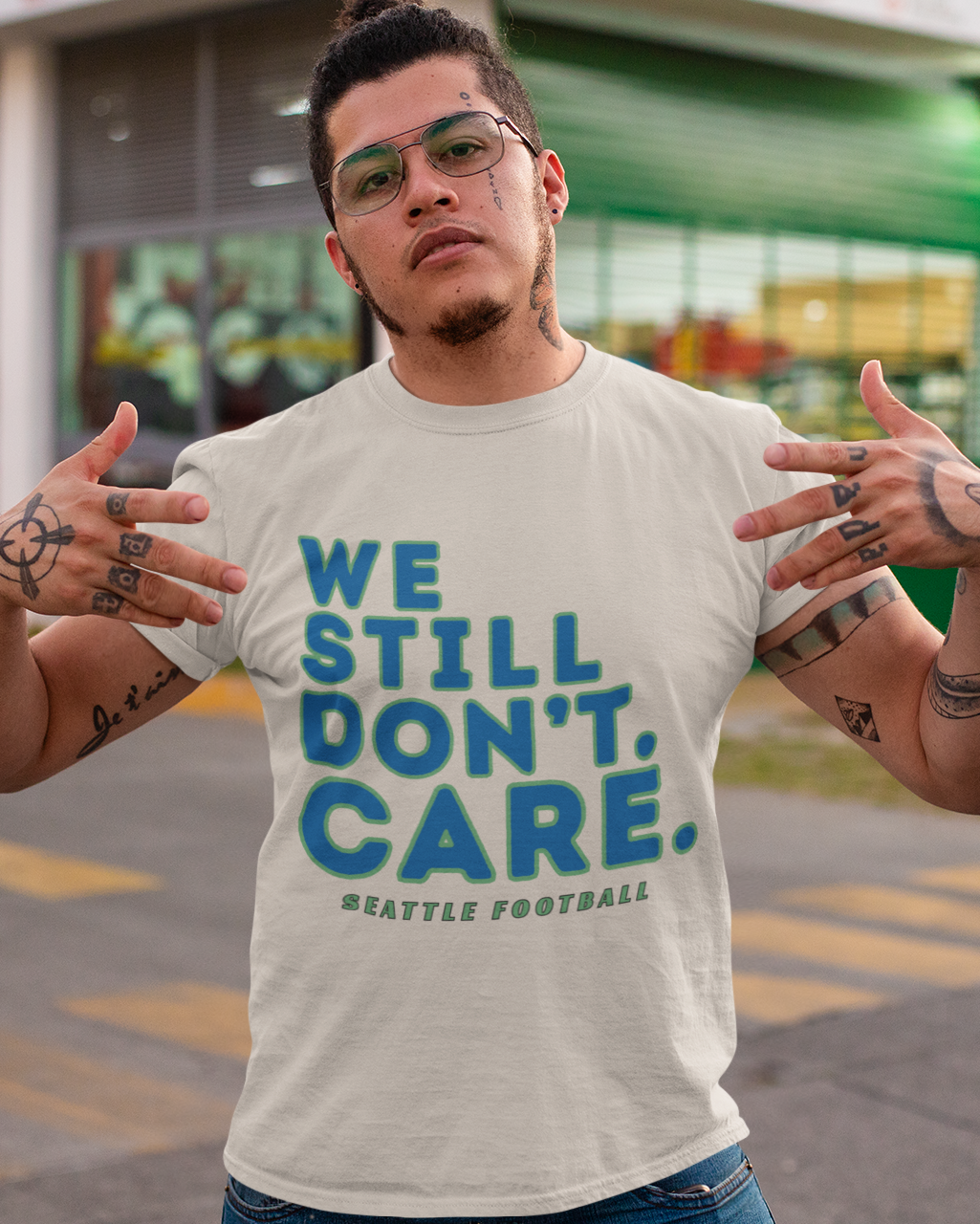 A man wearing a light beige graphic t-shirt with the text 'WE STILL DON'T CARE. SEATTLE FOOTBALL' in blue and green, posing outdoors in front of a storefront.