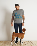 A man wearing a gray graphic t-shirt with blue text that reads 'WE STILL DON'T CARE.' stands next to a brown and white dog on a light wood floor against a white wall.
