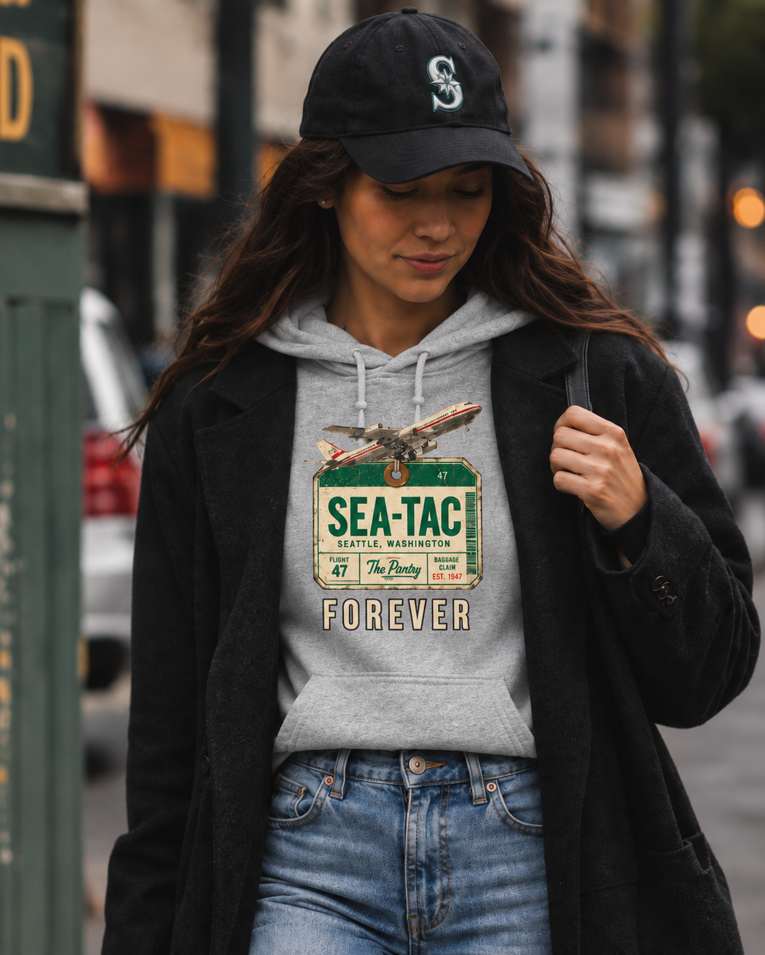 A woman walking on a city street wearing a gray hoodie with a vintage-style 'Sea-Tac' graphic, a black baseball cap, and a black coat.