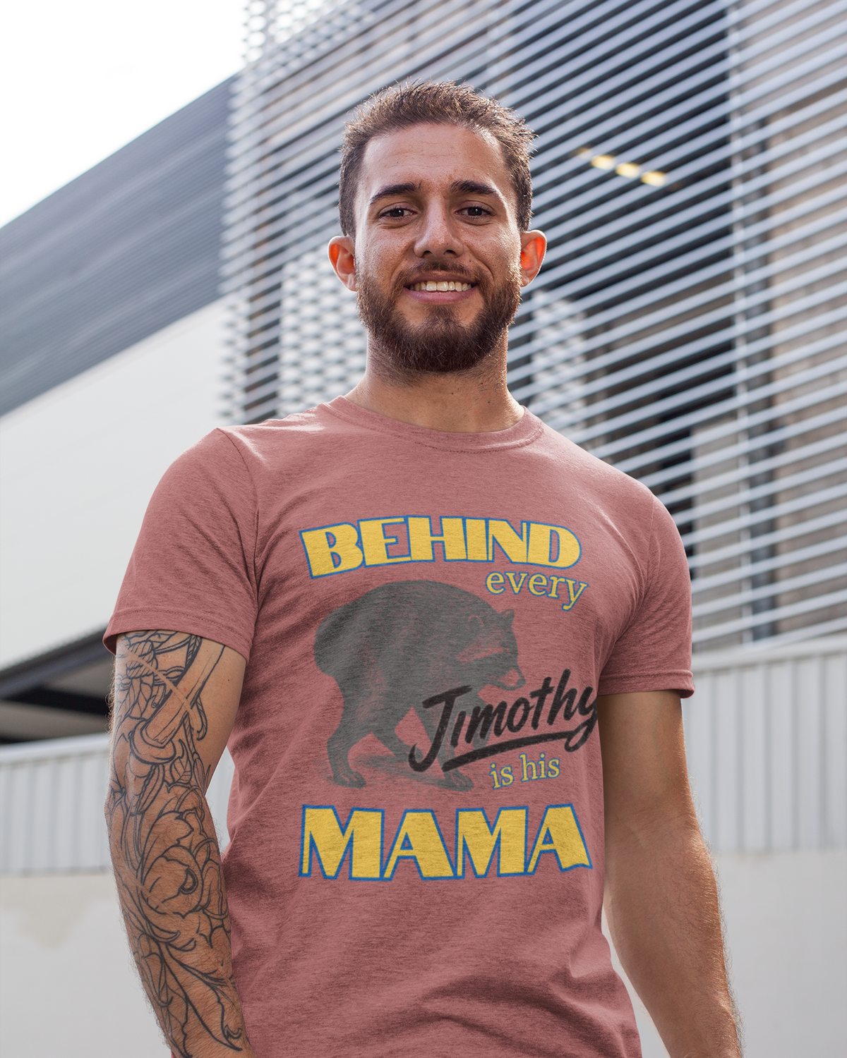 A man wearing a heathered dusty rose graphic t-shirt with Jimothy and the text 'BEHIND every Jimothy is his MAMA', paired with blue jeans, standing outdoors in front of a modern slatted building wall.