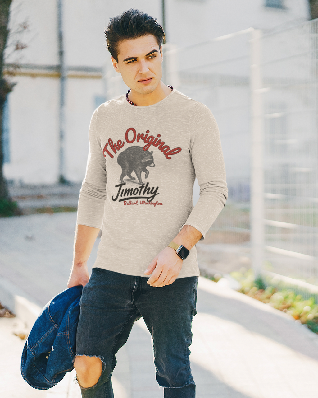 A man wearing a tan long-sleeve graphic sweatshirt with 'The Original Jimothy' and a raccoon print, paired with ripped dark jeans, walking outdoors next to a white wire fence.