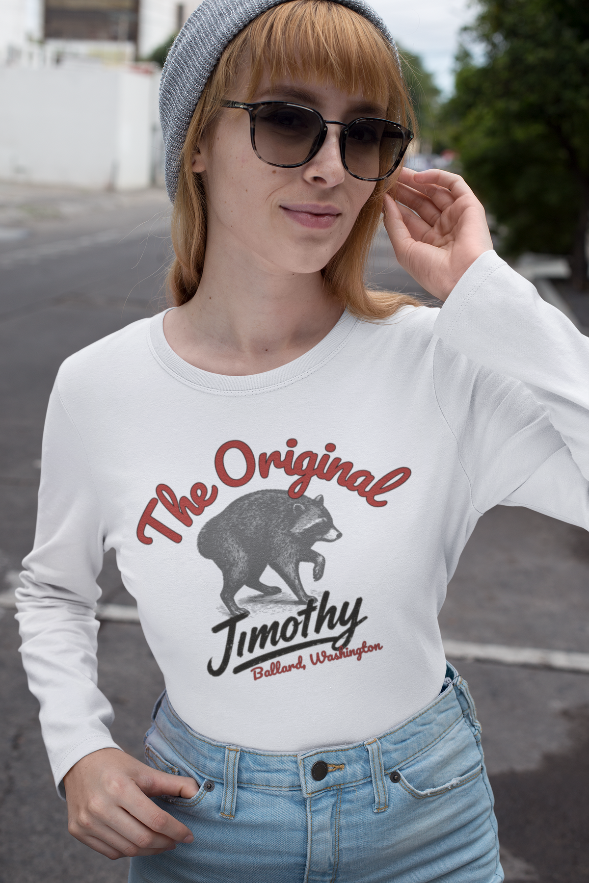 A woman wearing a white long-sleeve graphic t-shirt with a raccoon and the text 'The Original Jimothy' in red and black, paired with light blue jeans and a gray beanie, standing outdoors on a street.