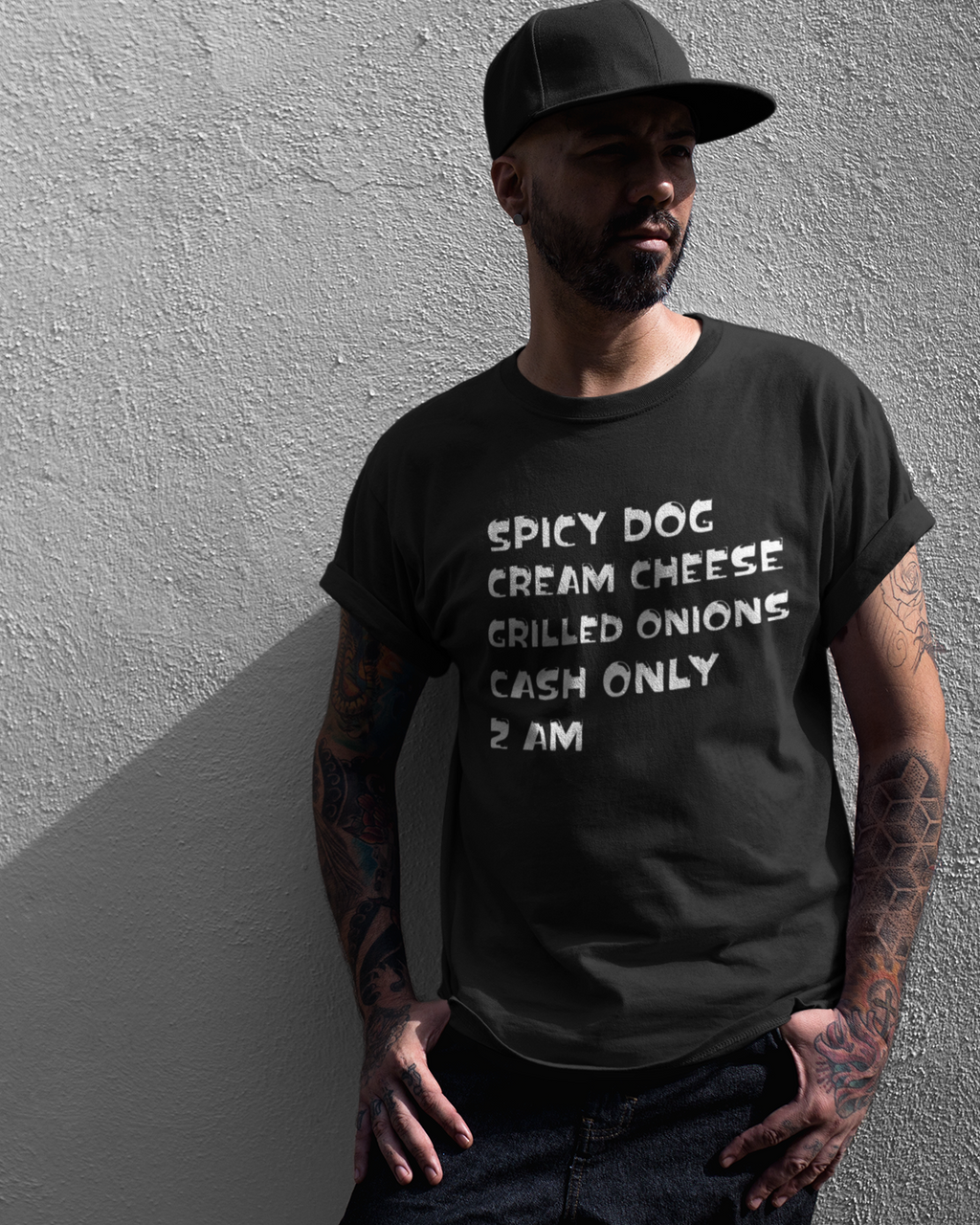 A man wearing a black graphic t-shirt with white text that reads 'SPICY DOG CREAM CHEESE GRILLED ONIONS CASH ONLY 2 AM' stands against a textured gray wall.