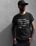 A man wearing a black graphic t-shirt with white text that reads 'SPICY DOG CREAM CHEESE GRILLED ONIONS CASH ONLY 2 AM' stands against a textured gray wall.