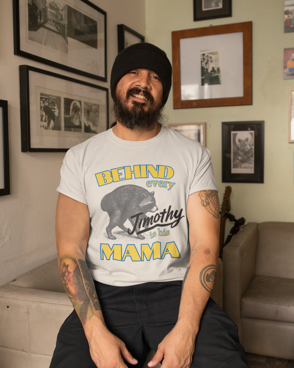 A man wearing a black beanie and a graphic t-shirt sitting in a living room. The t-shirt is light gray with a graphic of a bear and the text 'BEHIND every Jimothy and his MAMA'.