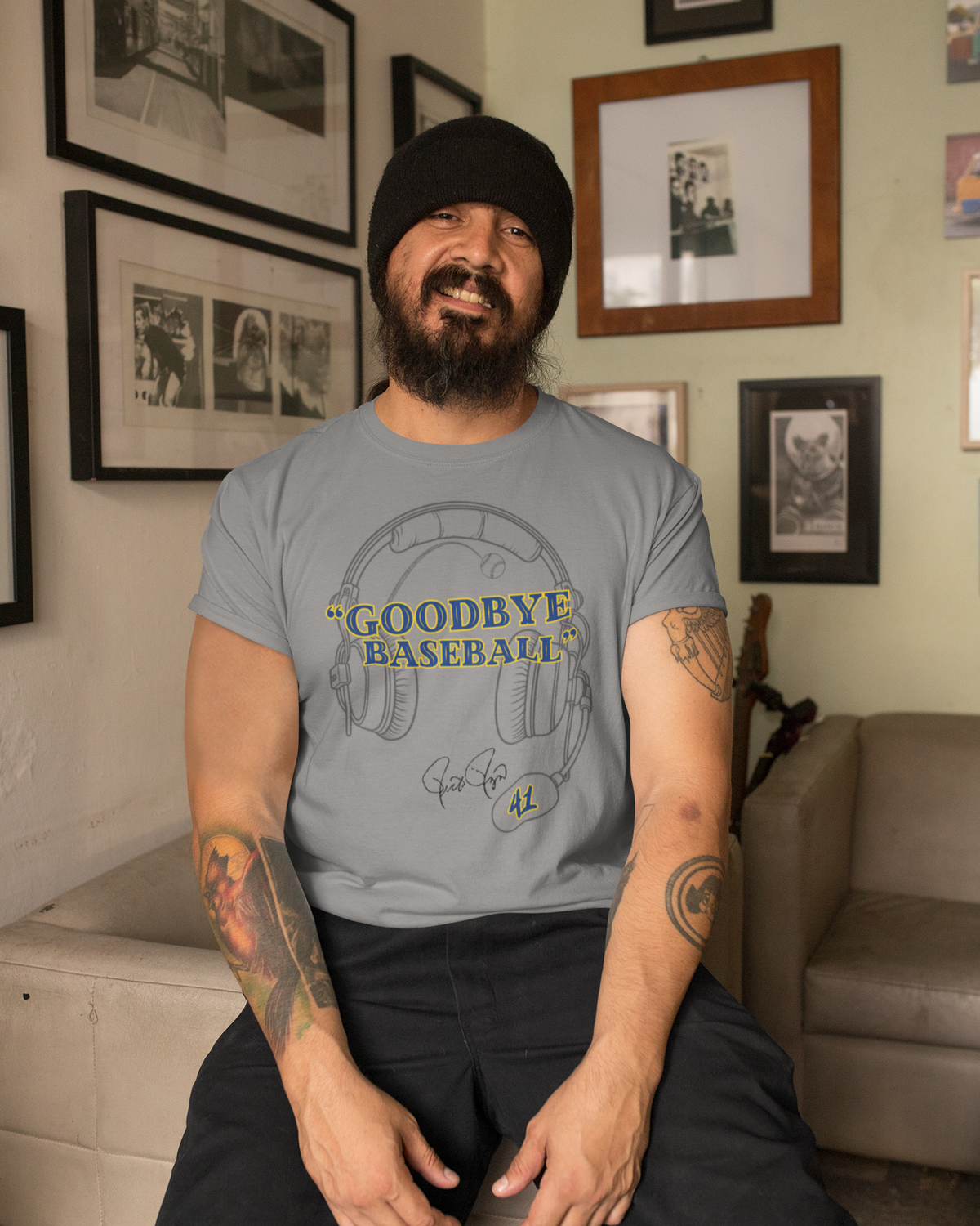 A man wearing a light gray graphic t-shirt with 'GOODBYE BASEBALL' and a baseball cap, sitting in a room with framed pictures on the wall.