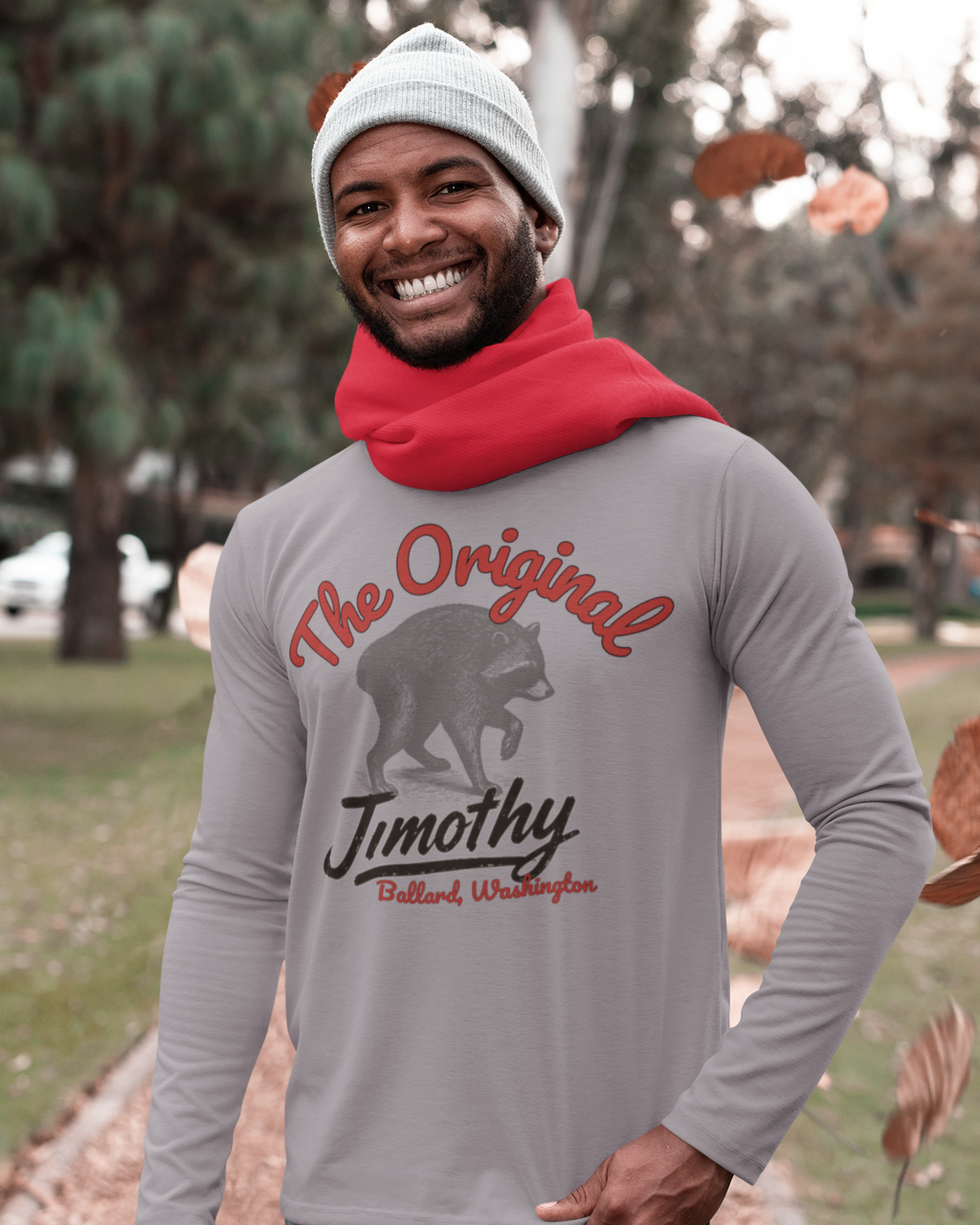 A man wearing a light gray long-sleeve graphic shirt with a red scarf and a red standing outdoor in a park. The shirt features a raccoon graphic and the text 'The Original Jimothy Ballard, Washington'.