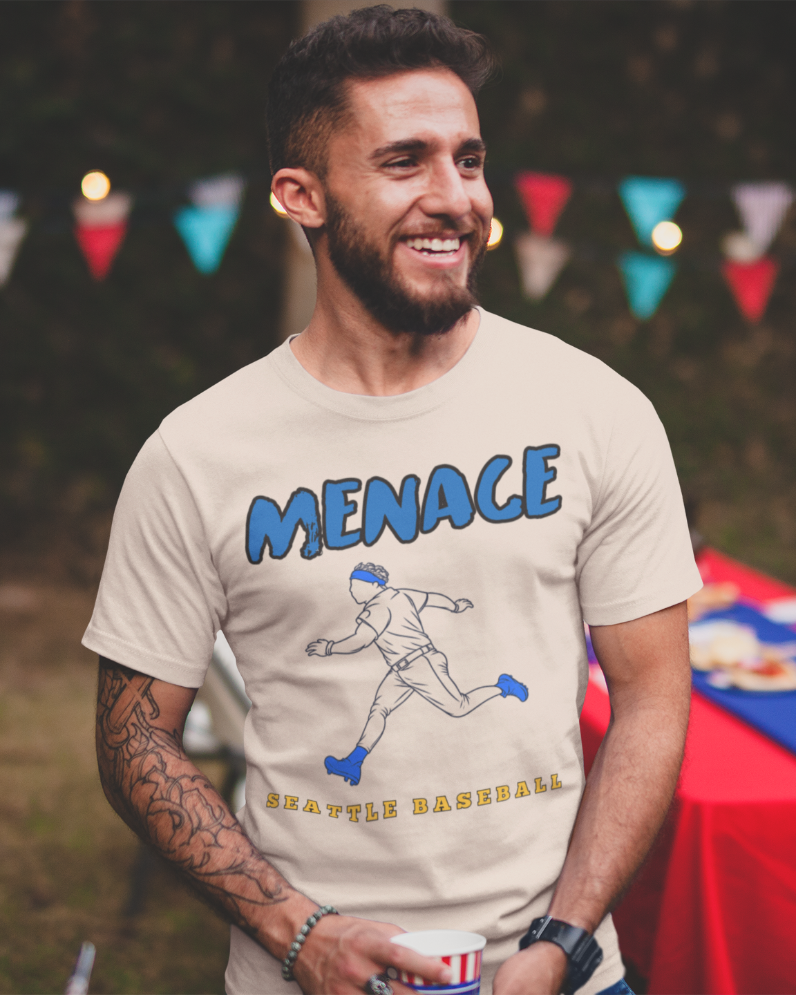 A smiling man at an outdoor party wearing a beige graphic t-shirt with 'MENACE' and a baseball player illustration in blue and yellow.