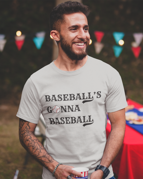 Man wearing a t-shirt with 'Baseball's Gonna Baseball' text at an outdoor event with string lights and a tablecloth.