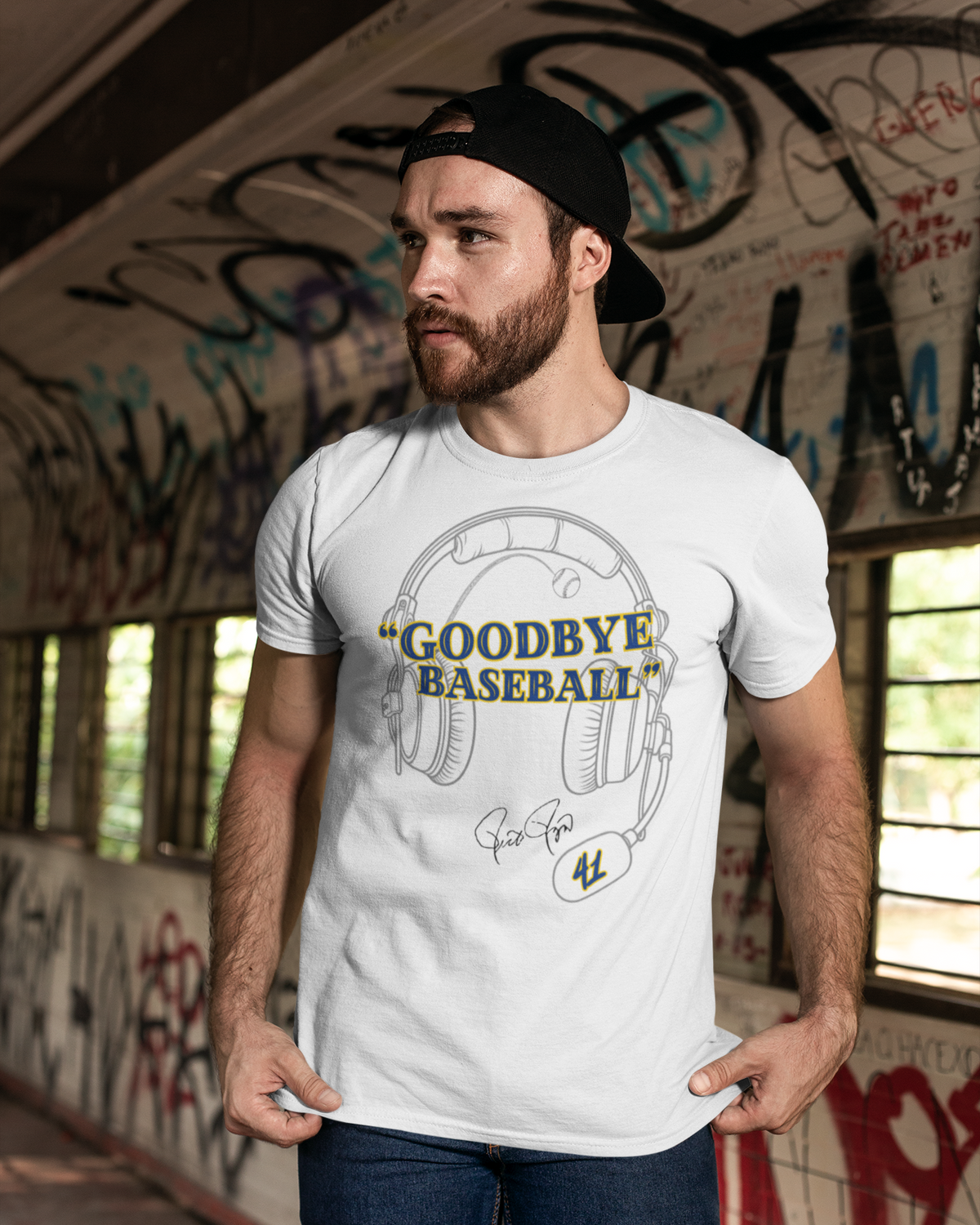 A man wearing a white graphic t-shirt with a baseball headset and 'GOODBYE BASEBALL' print, paired with dark blue jeans and a black cap, standing in a graffiti-covered corridor.