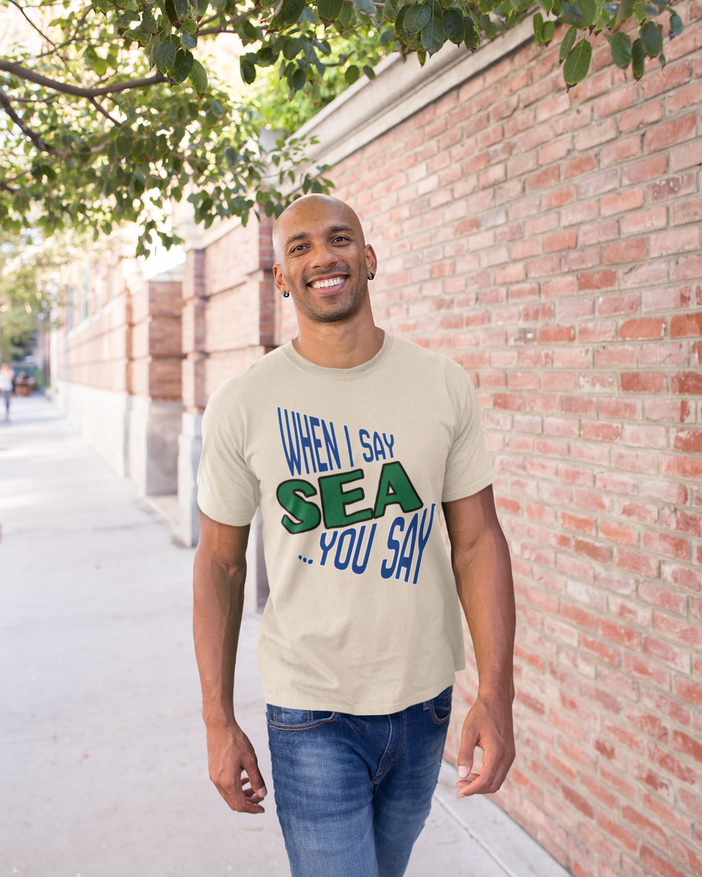 A man wearing a beige graphic t-shirt with blue and green text that reads 'WHEN I SAY SEA .