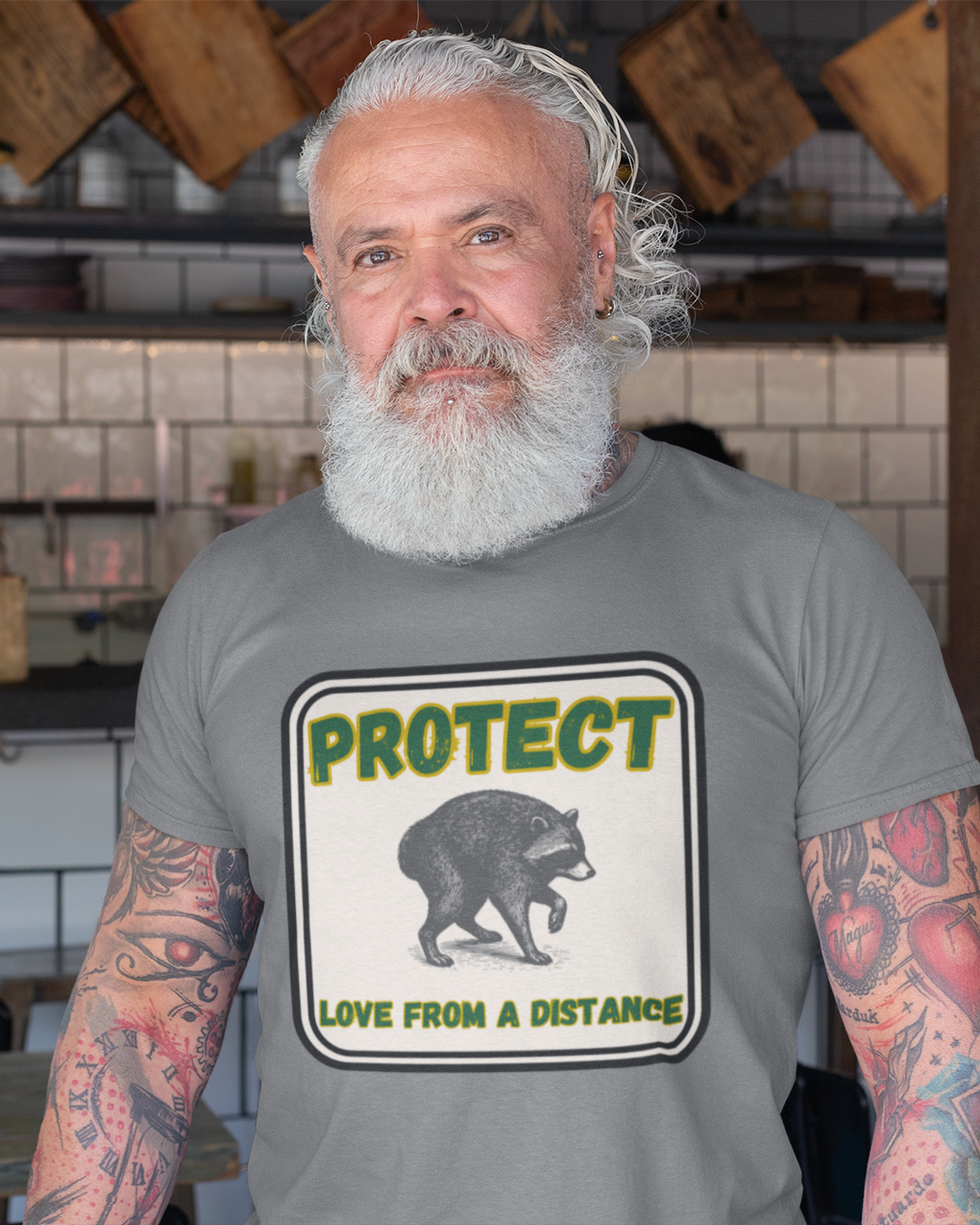 A man with a white beard and tattoos wearing a gray t-shirt with a 'PROTECT LOVE FROM A DISTANCE' bear graphic, standing in a bakery shop.