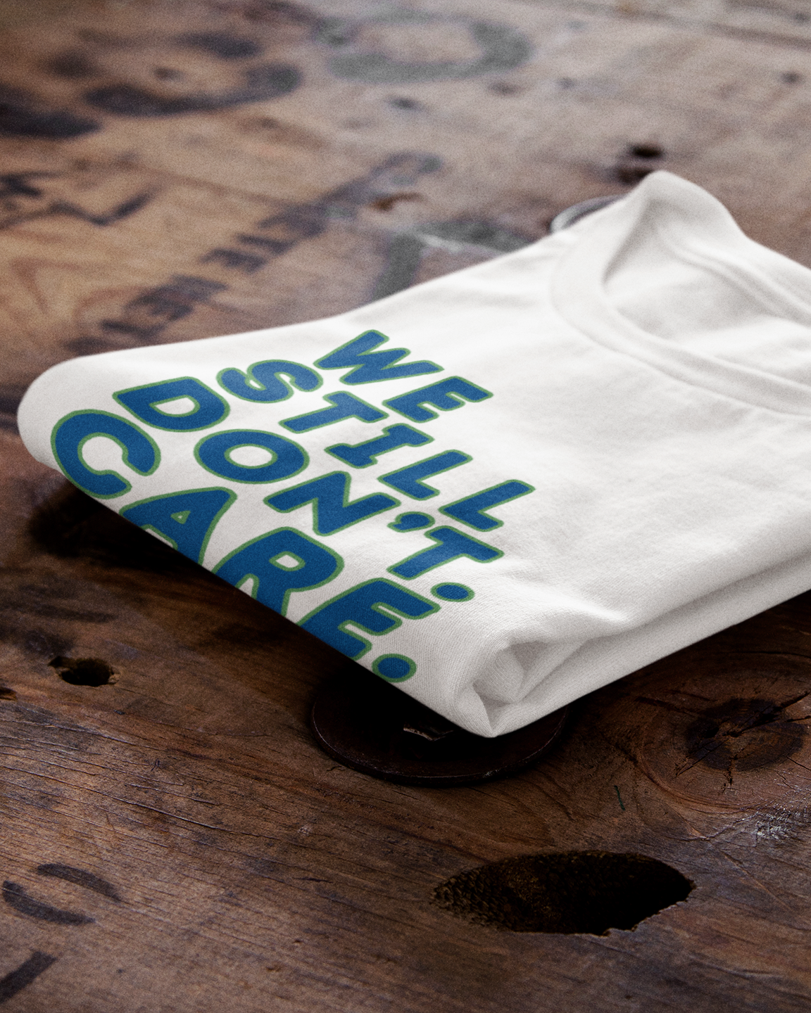 A folded white t-shirt with the text 'WE STILL DON'T CARE' printed in blue, displayed on a rustic wooden surface.