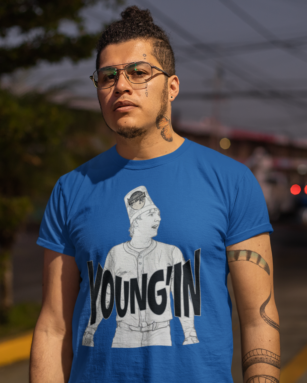 A man wearing a bright blue graphic t-shirt with a black and white illustration and the text 'YOUNG'IN' printed on the front, standing outdoors on a street at dusk.