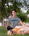 A smiling man in a gray graphic t-shirt lies on his back on a grassy lawn, playing with a golden retriever dog. The t-shirt features the word 'MENACE' in blue with a black-and-white graphic of a running figure.
