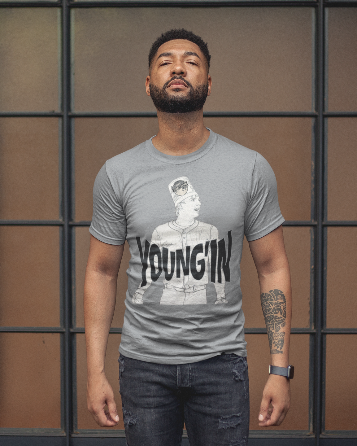 A man wearing a gray crewneck t-shirt with a black and white graphic print of a man's face and the word 'YOUNGIN' in bold letters, paired with dark blue jeans, standing in front of a brown window pane background.