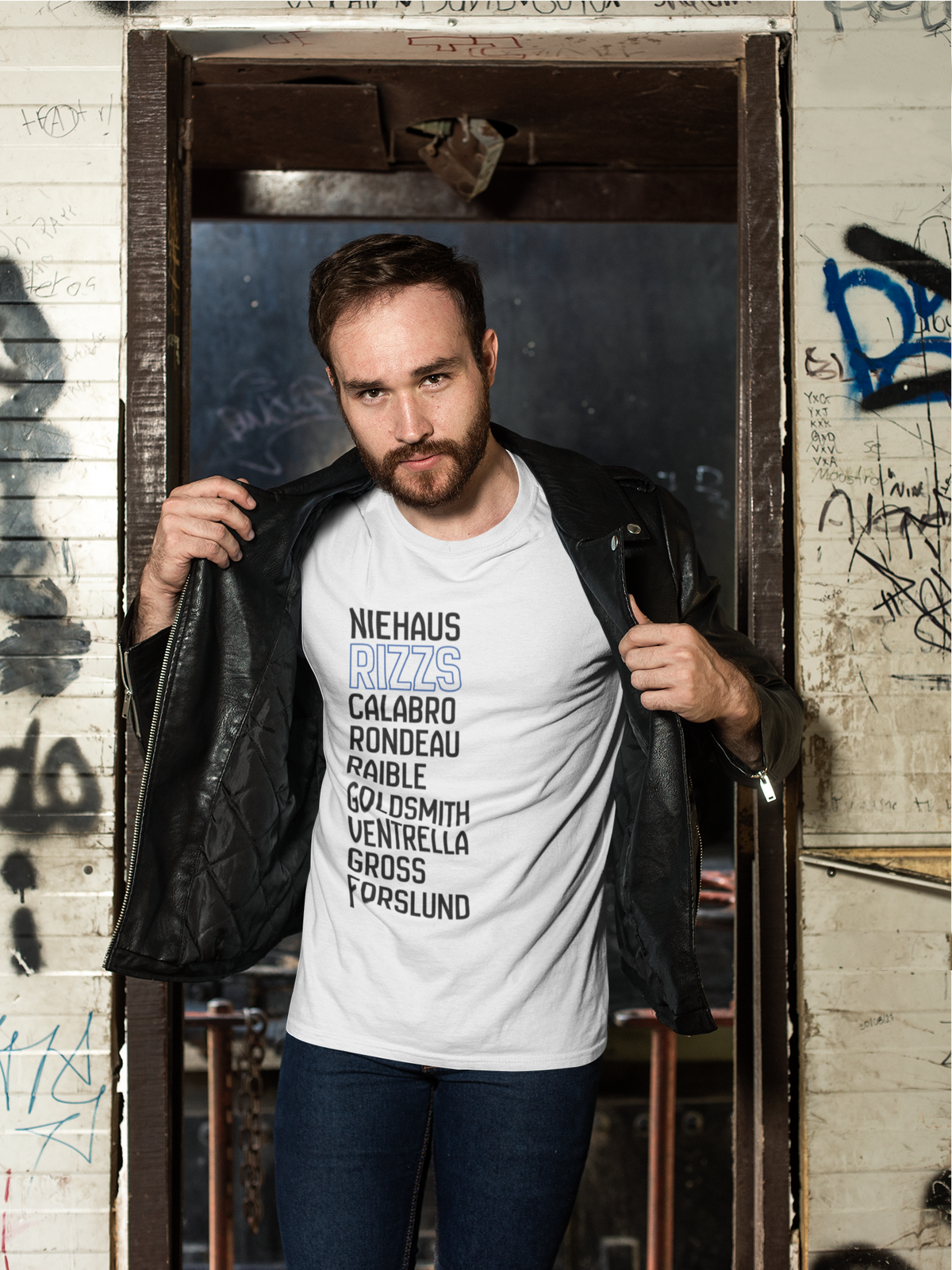 A man wearing a white graphic t-shirt with a list of names printed in black and light blue, styled with a black leather jacket and blue jeans, standing in a doorway of a graffiti-covered building.