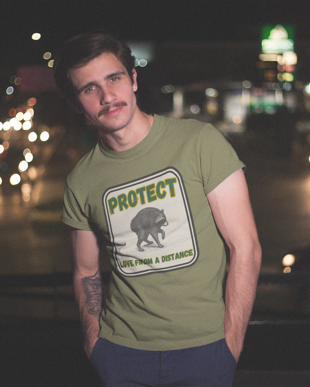A man wearing an olive green graphic t-shirt with a bear design and the text 'PROTECT ANIMALS A DISTANCE' stands in front of a blurred night street scene with car lights.