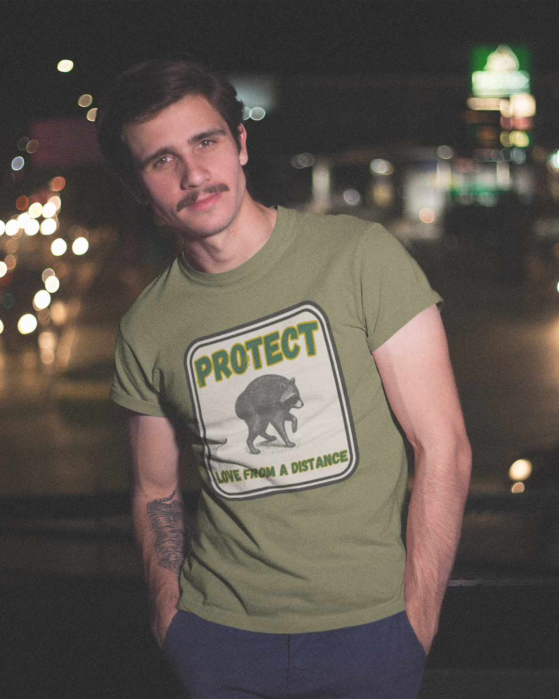 A man wearing an olive green graphic t-shirt with a bear design and the text 'PROTECT ANIMALS A DISTANCE' stands in front of a blurred night street scene with car lights.